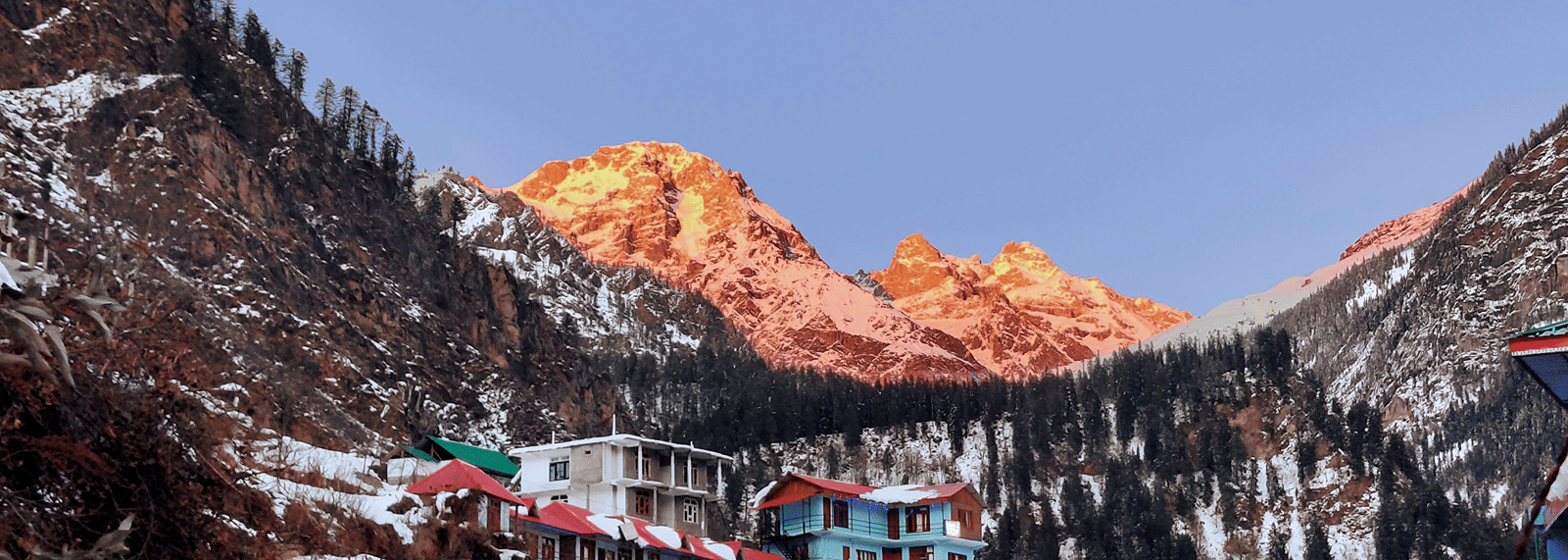 A high-angle view of houses nestled at the base of steep, snow-covered mountains, with sunlight illuminating the distant peaks during winter.