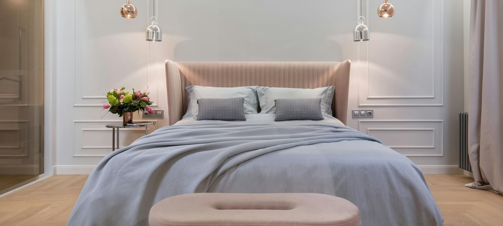 A modern bedroom is shown with soft lighting, a grey duvet, and a tufted bench.