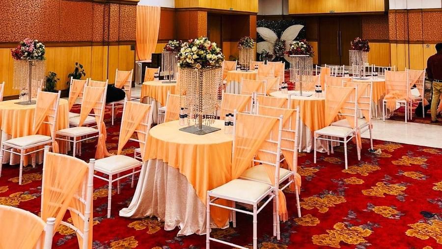  A banquet hall with a patterned carpet, set up with multiple round tables covered with table cloths and chairs.