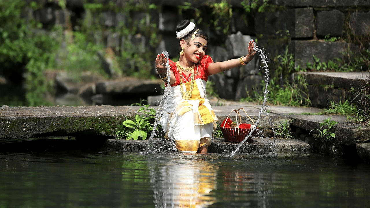 A girl in traditional Kerala attire performing a ritual near a serene water body.