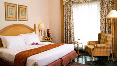 Elegant room at Savoy Suites, Noida with wooden headboard, plaid armchair, floral curtains, and warm traditional decor