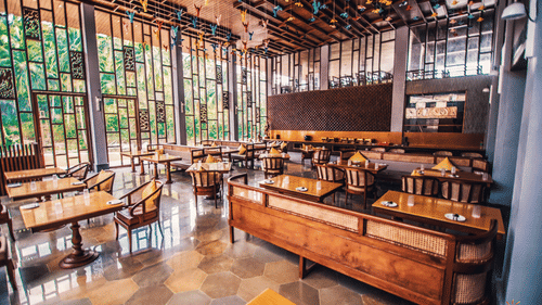 Daytime view of the restaurant at Symphony Oceanus Bay Beach Resort & Spa with wooden tables and cane chairs, a buffet counter, hexagonal tile floor, and glass walls facing greenery.