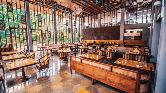Daytime view of the restaurant at Symphony Oceanus Bay Beach Resort & Spa with wooden tables and cane chairs, a buffet counter, hexagonal tile floor, and glass walls facing greenery.