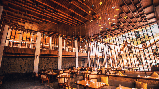 Wide angle of the restaurant at Symphony Oceanus Bay Beach Resort & Spa showing exposed wood beam ceiling, hanging bird ornaments, banquette seating, and a glazed facade opening onto the grounds.
