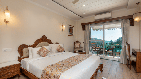  Hotel room at Quality Inn Dharamshala with a carved wooden king bed, floral bedspread, vintage chairs, wall-mounted air conditioner, and large curtained windows with garden views.