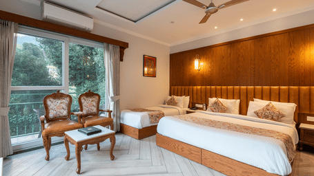 Deluxe room at Quality Inn Dharamshala with a king bed, wooden wall panelling, ceiling fan, vintage wooden chairs, and large windows overlooking lush garden greenery.