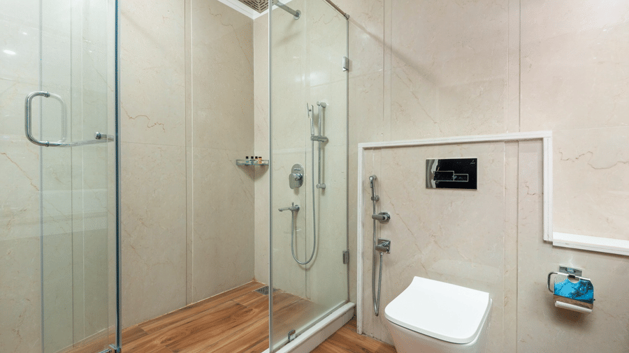 Modern bathroom at Quality Inn Dharamshala featuring a glass-enclosed shower cubicle with rainfall shower, wall-mounted toilet, and beige stone tiled walls.