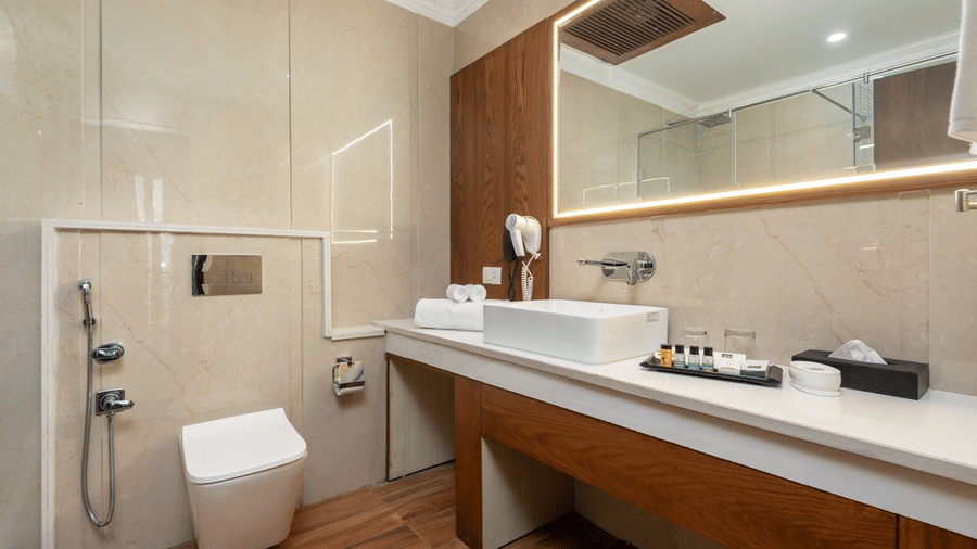 Spacious bathroom at Quality Inn Dharamshala with a long wooden vanity counter, vessel sink, large mirror, wall-mounted toilet, and warm beige tiled walls.