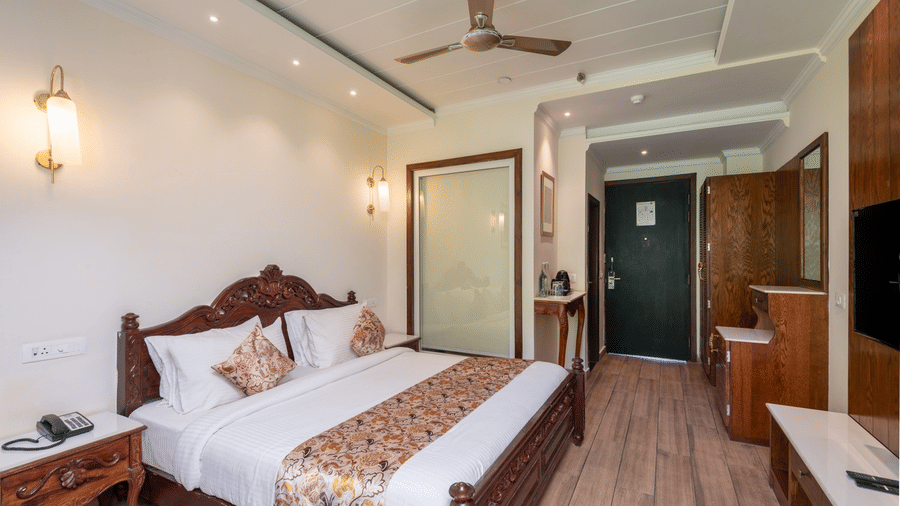 Wide angle view of a hotel room corridor at Quality Inn Dharamshala featuring twin carved wooden beds, floral bedspreads, wall sconces, ceiling fan, and room entrance door.