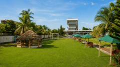 Long lawn at Pride Elite Mandarmani leading towards the hotel building, lined with a thatched gazebo, seating benches, and green sun umbrellas.
