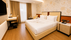 Deluxe Room bedroom view at Pride Premier Greater Noida showing a double bed, a bedside cabinet, a writing desk with a chair, and window curtains.