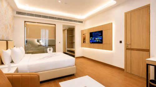 Suite bedroom view featuring a double bed, a wall-mounted television, wooden flooring, and a doorway at Pride Premier Greater Noida.