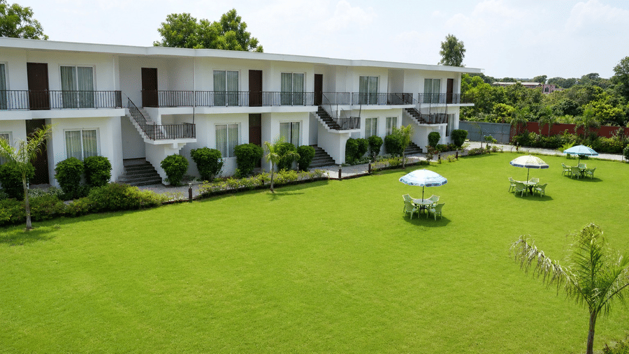 Exterior of a white 2-storey resort building with balcony railings and rooms facing a large manicured green lawn with shaded outdoor seating tables - Five Elements Hotels, Corbett Crown Resort, Jim Corbett