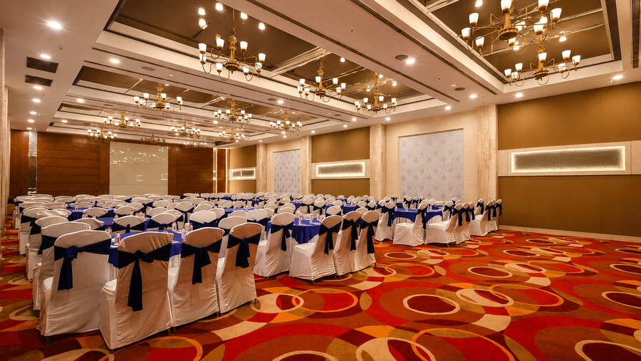 Large ballroom with blue-sashed white chairs, gold chandeliers, red patterned carpet, and classroom layout.