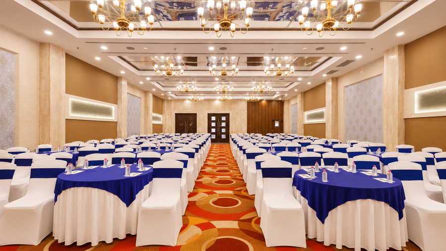 Grand ballroom with blue linen round tables, gold chandeliers, mirrored ceiling, and theatre-style setup.