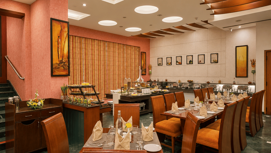 Spacious hotel dining area with wooden furniture, buffet counters, striped drapes, and vibrant artwork.