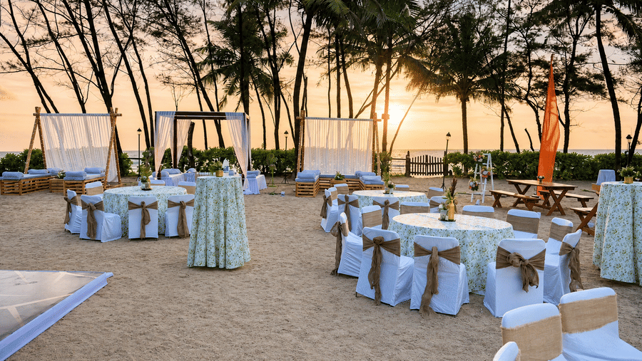 Outdoor beachfront event setup with decorated dining tables under palm trees at sunset at Kenilworth Resort & Spa, Goa.