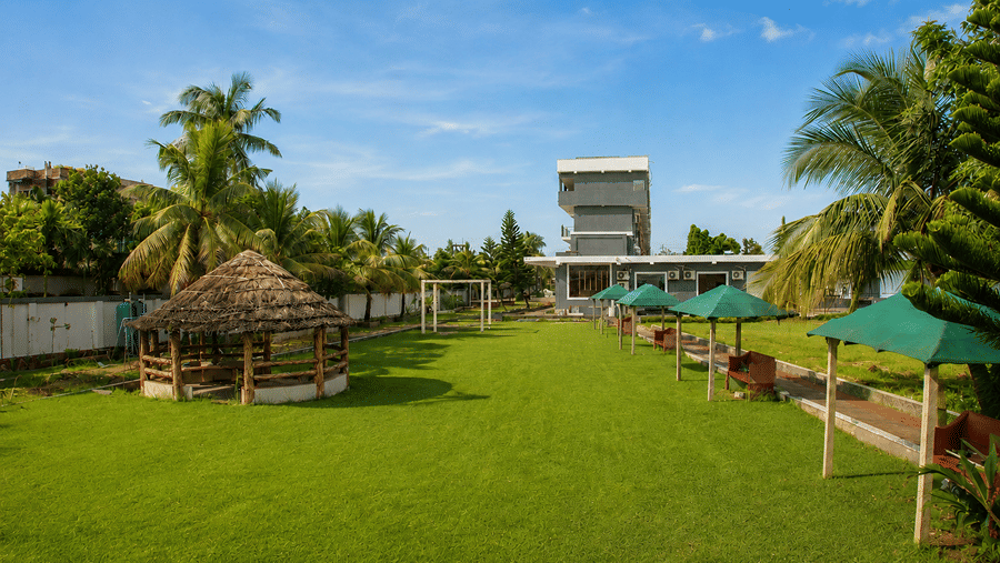 Long lawn at Pride Elite Mandarmani leading towards the hotel building, lined with a thatched gazebo, seating benches, and green sun umbrellas.