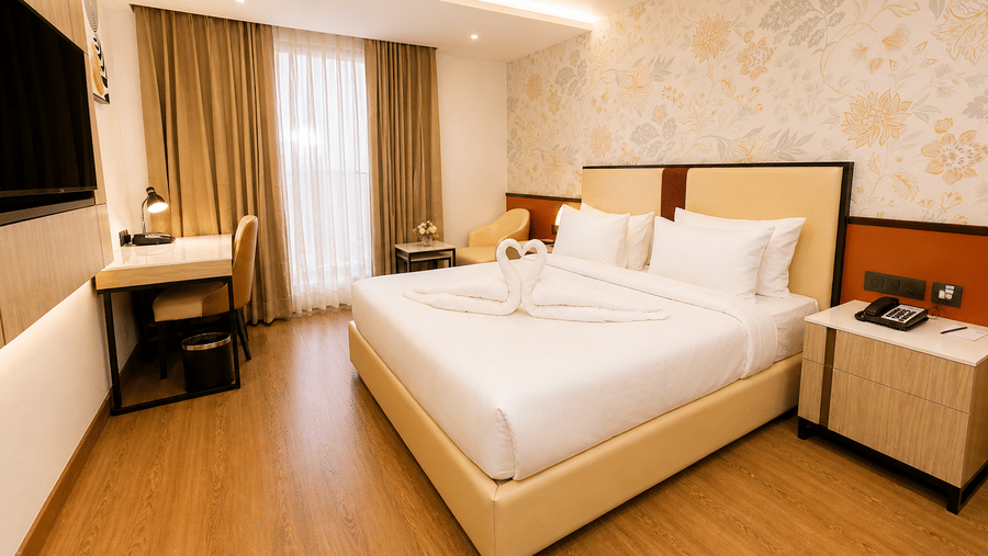 Deluxe Room bedroom view at Pride Premier Greater Noida showing a double bed, a bedside cabinet, a writing desk with a chair, and window curtains.