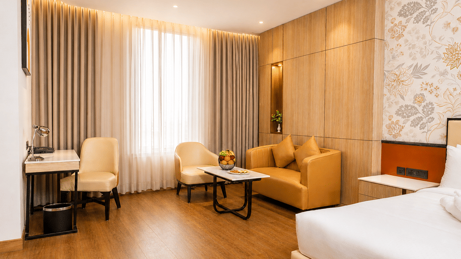 A spacious Premium Room layout at Pride Premier Greater Noida featuring a seating area with a sofa, armchairs, a coffee table, and a bed in the foreground.