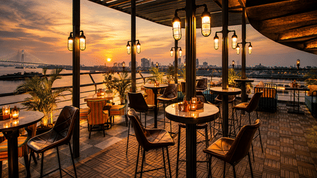 An outdoor rooftop bar or restaurant at sunset, featuring high-top tables and dark bar stools under a covered pergola.