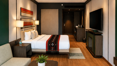 Hotel bedroom featuring double bed with decorative runner, wall-mounted television, sofa, and wooden flooring.