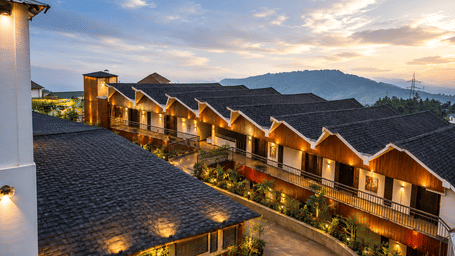 High-angle twilight view of hotel tiled roofs with warm exterior lighting, walkway, and distant mountains under dusk sky.