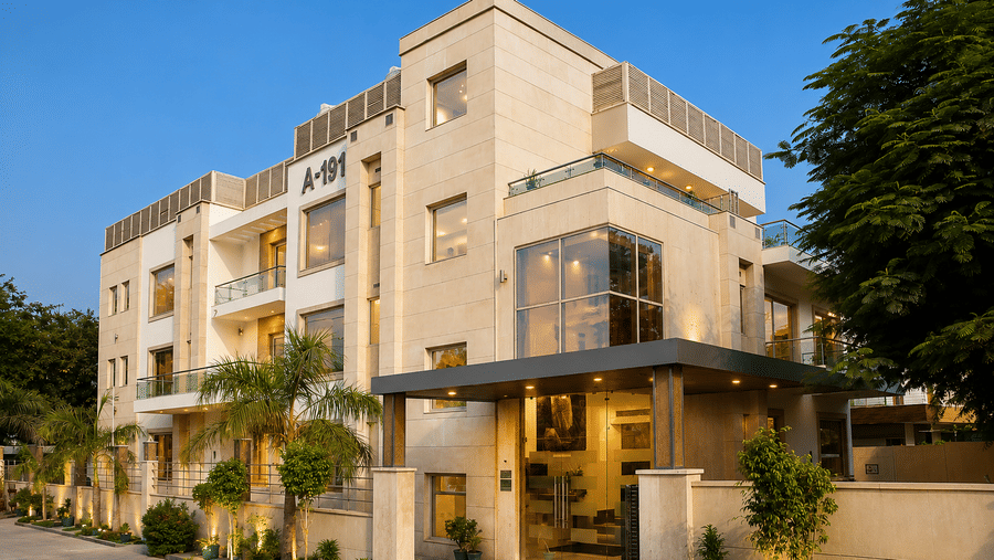 Hotel building exterior in daylight with a glass canopy entrance, tall trees and a boundary wall running along the driveway close by.