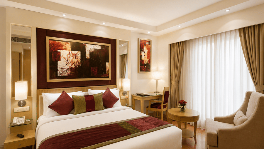 Hotel room with a bed dressed in white and maroon linen, framed wall artwork, a beige armchair and long green curtains by the window nearby.