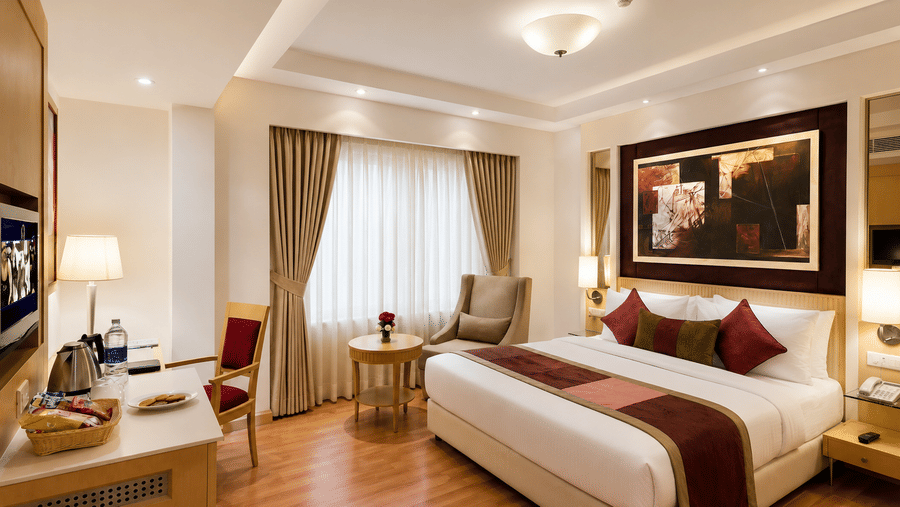 Hotel room with a bed dressed in white and maroon linen, framed wall artwork, a beige armchair and warm wooden flooring throughout the room.