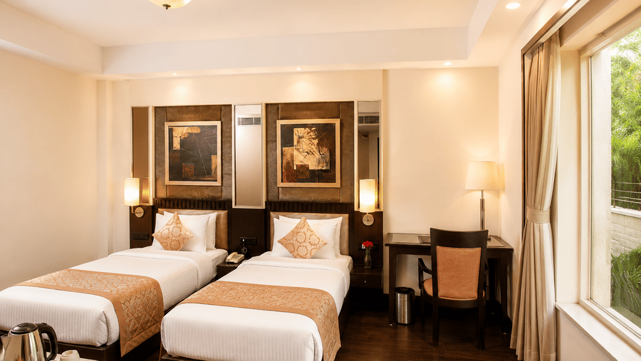 Hotel room with twin beds dressed in white linen, framed wall artwork, a work desk and long curtains by a window overlooking green trees.