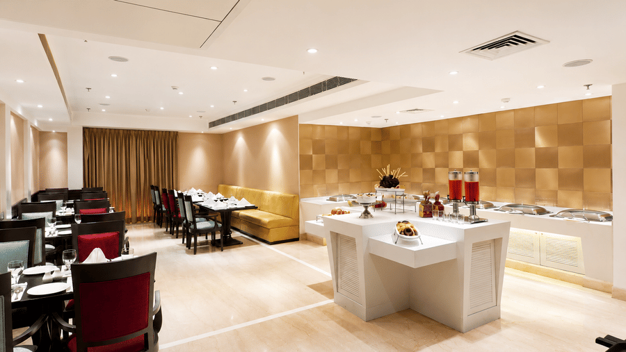 Restaurant seating area with red upholstered chairs, dark wooden tables and a buffet counter set against warm ambient lighting throughout.