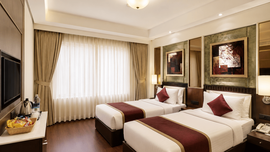 Hotel room with twin beds dressed in white and maroon linen, framed wall artwork, a wall-mounted television and long sheer curtains nearby.