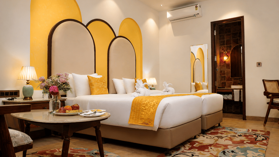 Wide view of a guest room with an arched headboard, double bed, yellow accents, and a floral carpet at Kadamb Kuteer, Vrindavan.