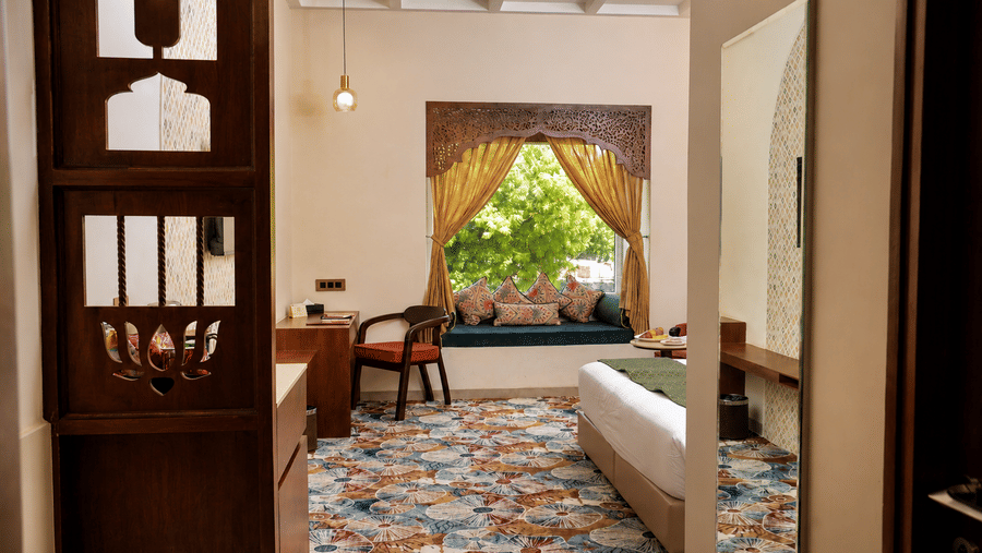 View through a wooden doorway into a guest room with a patterned floor and garden beyond at Kadamb Kuteer, Vrindavan.