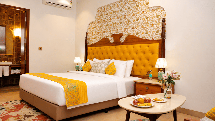 Guest room with a golden tufted headboard, yellow bed runner, and a side table with a food tray at Kadamb Kuteer, Vrindavan.