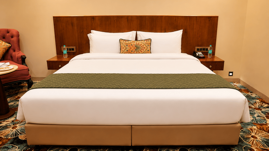 King bed with a wooden headboard, white bedding, and a vibrant patterned carpet at Kadamb Kuteer, Vrindavan.