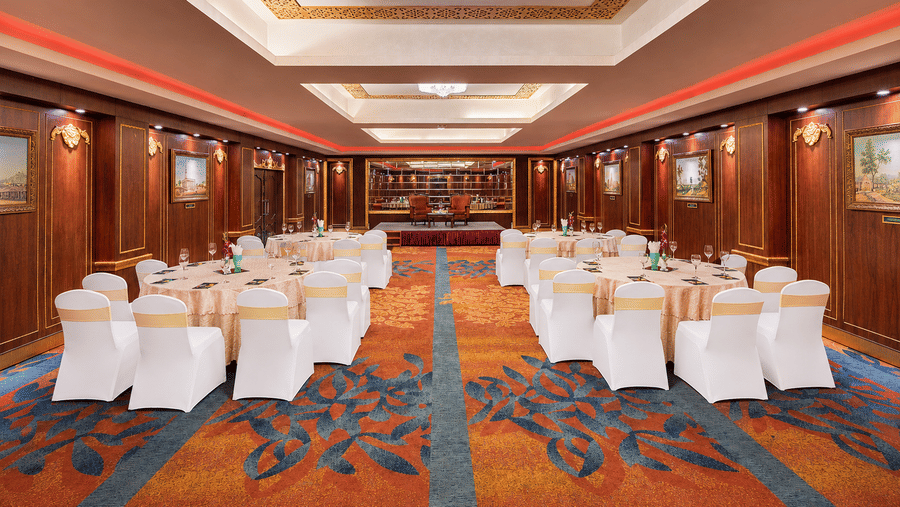 Tulip banquet hall with round tables, white chair covers, patterned carpet, wood-panelled walls with framed paintings and a stage at MAYFAIR Garden, Rourkela.