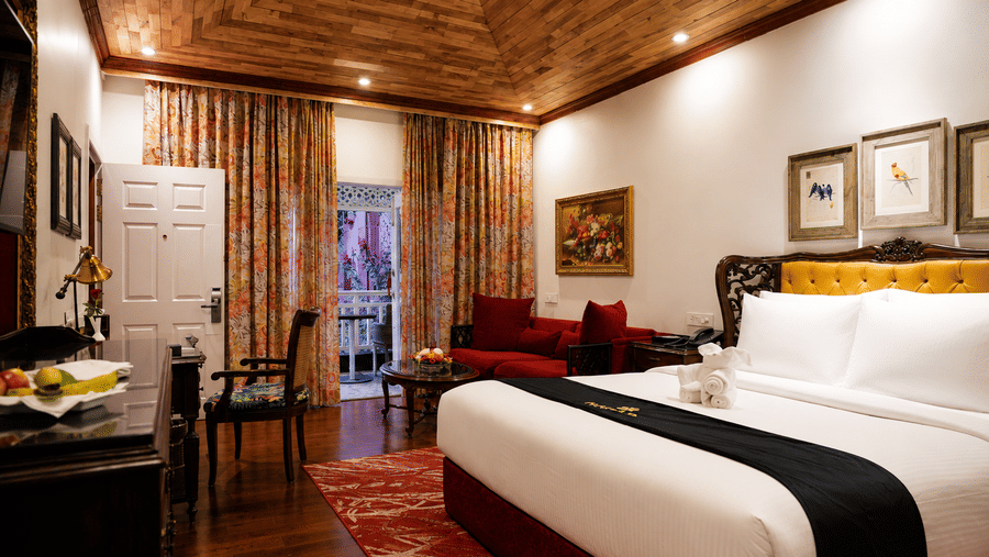 Deluxe Cottage at MAYFAIR Himalayan Spa Resort Kalimpong featuring a bed in the foreground. A sofa sits near a door that opens to the outside. Framed pictures hang on the walls. The ceiling is clad in wood.