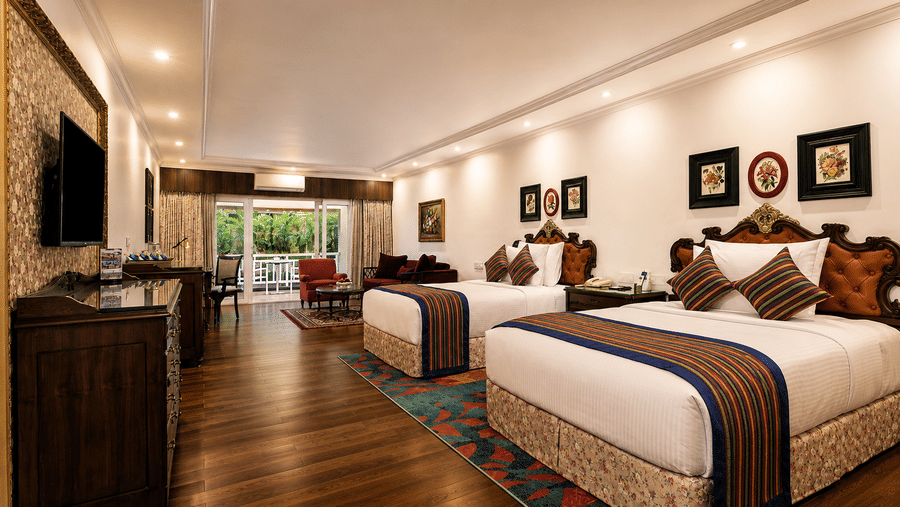Executive Room at MAYFAIR Himalayan Spa Resort Kalimpong showing two beds with patterned runners. A television is mounted on the left wall near a storage cabinet. An open doorway leads to another section of the property.