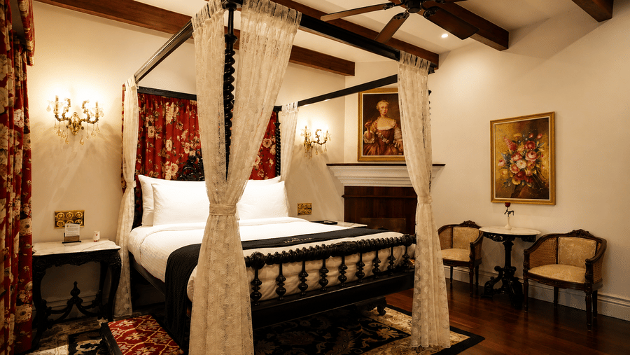 Heritage Room featuring a bed with a four-poster frame and curtains at MAYFAIR Himalayan Spa Resort Kalimpong. A ceiling fan hangs above. Two chairs and a small table sit near a fireplace. Framed artwork is mounted on the wall.
