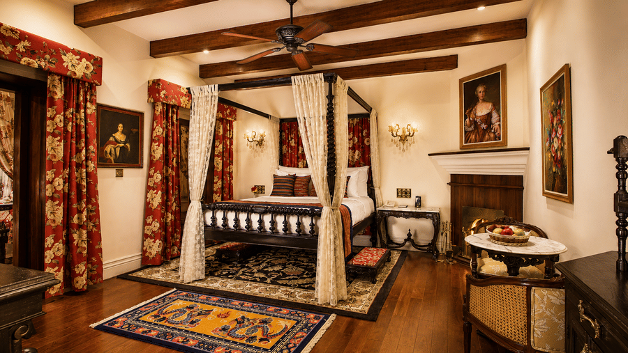 Heritage Suite dedroom at MAYFAIR Himalayan Spa Resort Kalimpong. A four-poster bed with curtains stands on a rug. Wooden beams line the ceiling. Paintings hang on the walls. A fireplace sits in the corner next to a table and chairs.