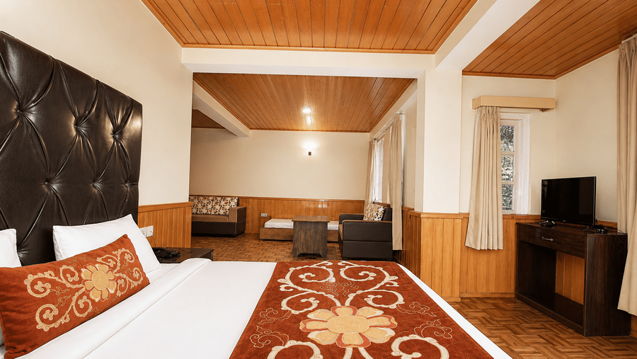 A bed with pillows occupies the foreground of the Junior Suite at Orsino Khangri Karpo Retreat & Spa, Lachung. A sofa and table sit in the background area. Timber panels line the ceiling.