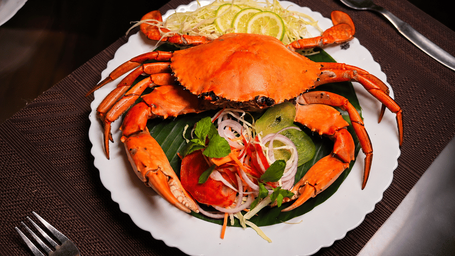 Overhead view of a plated crab dish garnished with lemon slices and fresh salad, served on a white plate with cutlery placed alongside it.