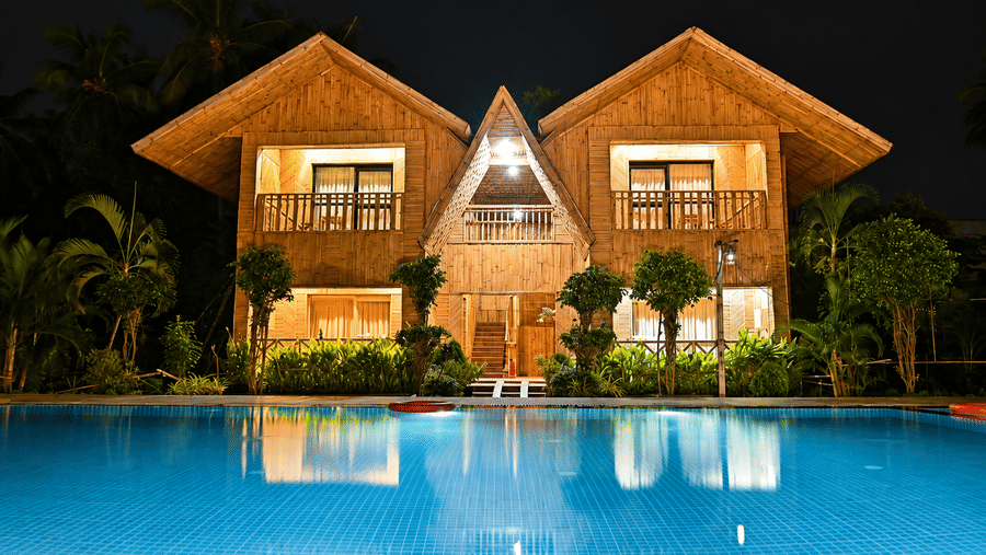 Resort building exterior at night with wooden facades, illuminated windows, palm trees and a swimming pool reflecting the lights in the front.