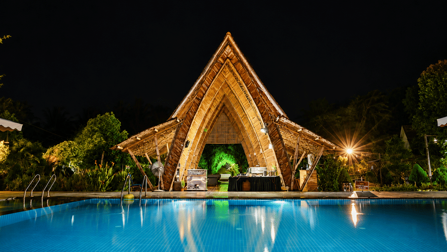 Triangular wooden pavilion lit up at night with green ambient lighting, palm trees around it and a swimming pool reflecting the lights below.