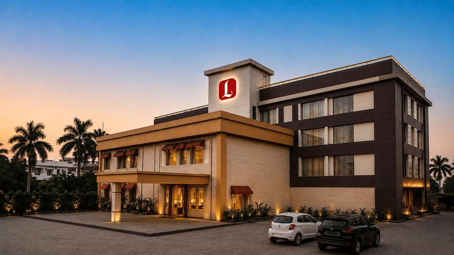 Exterior view of a modern multi-story hotel illuminated at dusk, featuring a paved parking area with parked cars and palm trees in the background - Rang Lords Inn Bharuch 