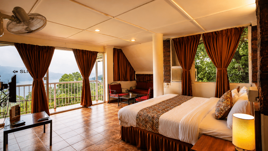 Wide-angle view of a Wind Chalet bedroom showing double bed, bedside lamp, fan, and open balcony doors with scenic view.