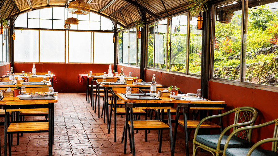 Covered terrace dining area at Wind Chalet featuring wooden tables, red brick tile flooring, and large glass windows.