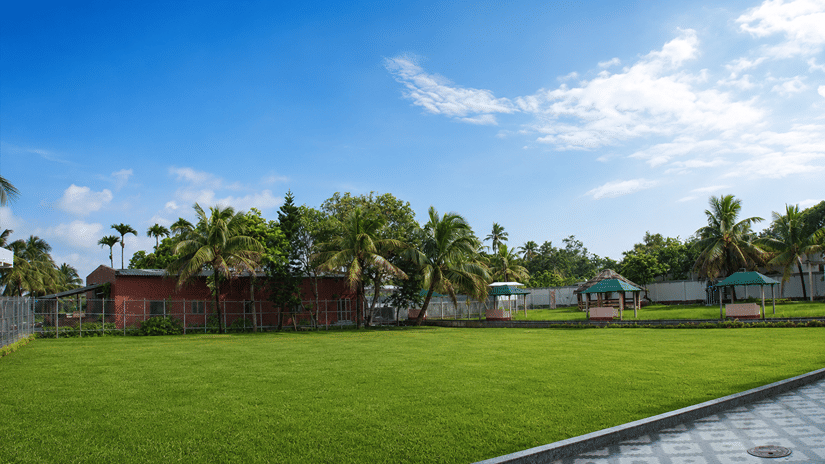 Pride Elite Mandarmani grounds with a manicured lawn, palm trees, a red building, and 2 green thatched-roof gazebos under a bright blue sky.
